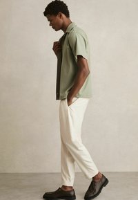 Camisa de manga corta, verde claro, con un diseño de botones, combinada con pantalones crema y zapatos marrón oscuro. Tejido suave y corte ajustado.