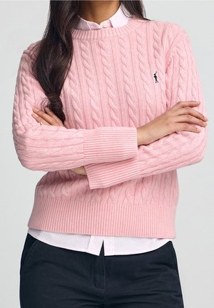 Femme aux longs cheveux foncés, portant un pull en maille torsadée rose sur une chemise rose clair à col, bras croisés, et pantalon foncé.