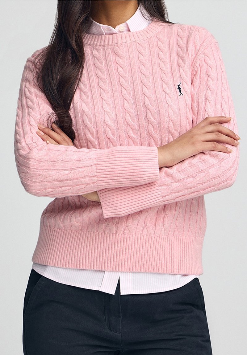 Femme aux longs cheveux foncés, portant un pull en maille torsadée rose sur une chemise rose clair à col, bras croisés, et pantalon foncé.