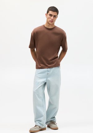 Bruine korte mouwen t-shirt, relaxte fit, gecombineerd met lichtblauwe baggy jeans en tan schoenen, tegen een witte achtergrond.