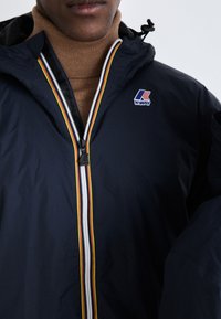 Veste coupe-vent navy avec une fermeture éclair à l'avant, dotée d'une rayure colorée et d'un patch logo. Col ajusté et tissu texturé.