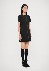 Černé krátké šaty s krátkým rukávem a přímým střihem, zdobené zlatými cvočky vpředu, doplněné o černé podkolenky a otevřené podpatky.