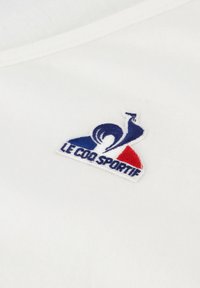 Bílý bavlněný tričko s vyšitým logem v modré, červené a bílé barvě, s nápisem "Le Coq Sportif." Hladká textura.