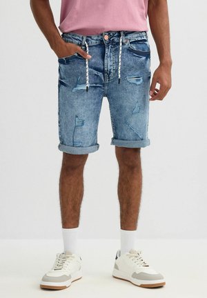 Hombre con pantalones cortos de mezclilla azul desgastados y remangados con cordón blanco, calcetines blancos y zapatillas blancas, de pie con una mano en el bolsillo.