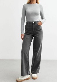 Harmaa pitkähihaisen paidan parina on korkeavyötäröiset, leveät harmaat farkut. Kengät ovat valkoiset, joissa on vaaleanruskeat pohjat. Minimalistinen muotoilu, rento ilme.