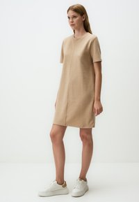 Beiges Kurzarmkleid mit geradem Schnitt, Rundhalsausschnitt und mittiger Nahtdetails. Kombiniert mit weißen Sneakern mit beigen Akzenten.