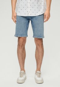 Shorts de mezclilla azul claro con pierna recta, costuras visibles y dobladillos enrollados, combinados con una camisa blanca con estampado y zapatillas.