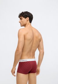 Ung mann med kort krøllete hår iført røde Calvin Klein boxershorts med et geometrisk mønster, vendt bort fra en ensfarget bakgrunn.