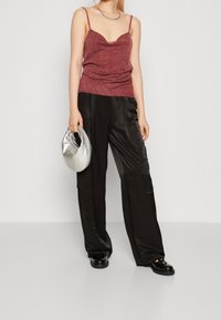 Donna che indossa un top senza maniche lucido color marrone scuro e pantaloni larghi in satin neri, con in mano una borsa rotonda argentata e indossando mocassini neri.