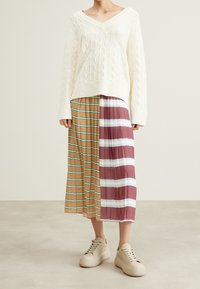 Pull en tricot blanc avec un col en V et un motif texturé, associé à une jupe plissée présentant des rayures marron, vert et bordeaux. Baskets beiges.