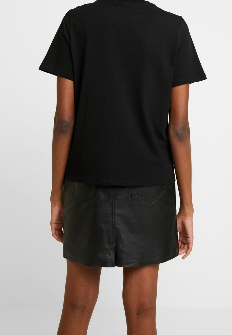 Une personne portant un t-shirt noir à manches courtes et une mini-jupe en cuir noire se tenant de dos face à la caméra.