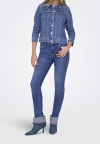 Denim jacket with button closure, two chest pockets, and a mid-blue wash; paired with fitted jeans featuring a cuffed hem and pointed heels.