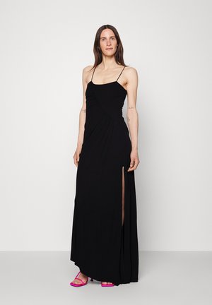 Kvinne iført en svart ermeløs kveldsnattkjole med sideslitt og rosa høyhælte sandaler, stående mot en enkel lys bakgrunn.