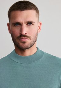 Lys teal sweater med en høj, ribbet krave. Stoffet fremst�år glat og tætsiddende, hvilket komplementerer personens ansigtstræk.