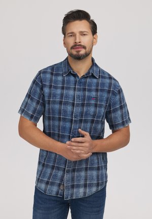 Mężczyzna z brodą w niebieską kratowaną koszulę z krótkim rękawem i dżinsami, stojący ze splecionymi rękami na jasnoszarym tle.