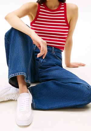 Personne assise portant un haut sans manches rayé rouge et blanc, un jean bleu à jambes larges et des baskets blanches sur un fond clair.