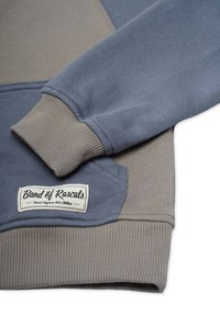 Harmaa ja sininen collegepaita, jossa on ribbatut hihansuut, pyöreä helma ja etiketti, jossa lukee "Band of Rascals". Pehmeää puuvillakangasta, tilkkutyösuunnittelu.