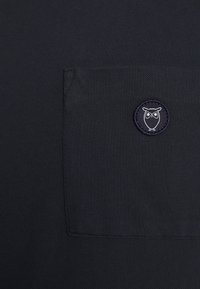 Marineblaue Baumwollbluse mit einer Brusttasche und einem runden Eulenlogo, das in Weiß auf marineblauem Hintergrund gestickt ist. Texturiert.