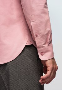 Main d'homme à côté d'un poignet de chemise rose à manches longues avec deux boutons, portée sur un pantalon à carreaux sombres sur un fond uni.