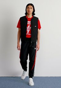 Colete acolchoado preto sobre uma t-shirt vermelha com logótipo, combinado com calças pretas com riscas vermelhas e brancas, e ténis brancos.