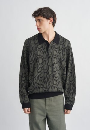 Giovane uomo rivolto verso sinistra che indossa una camicia a maniche lunghe con colletto nero e stampa grigia a motivo serpente, abbinata a pantaloni verde oliva.