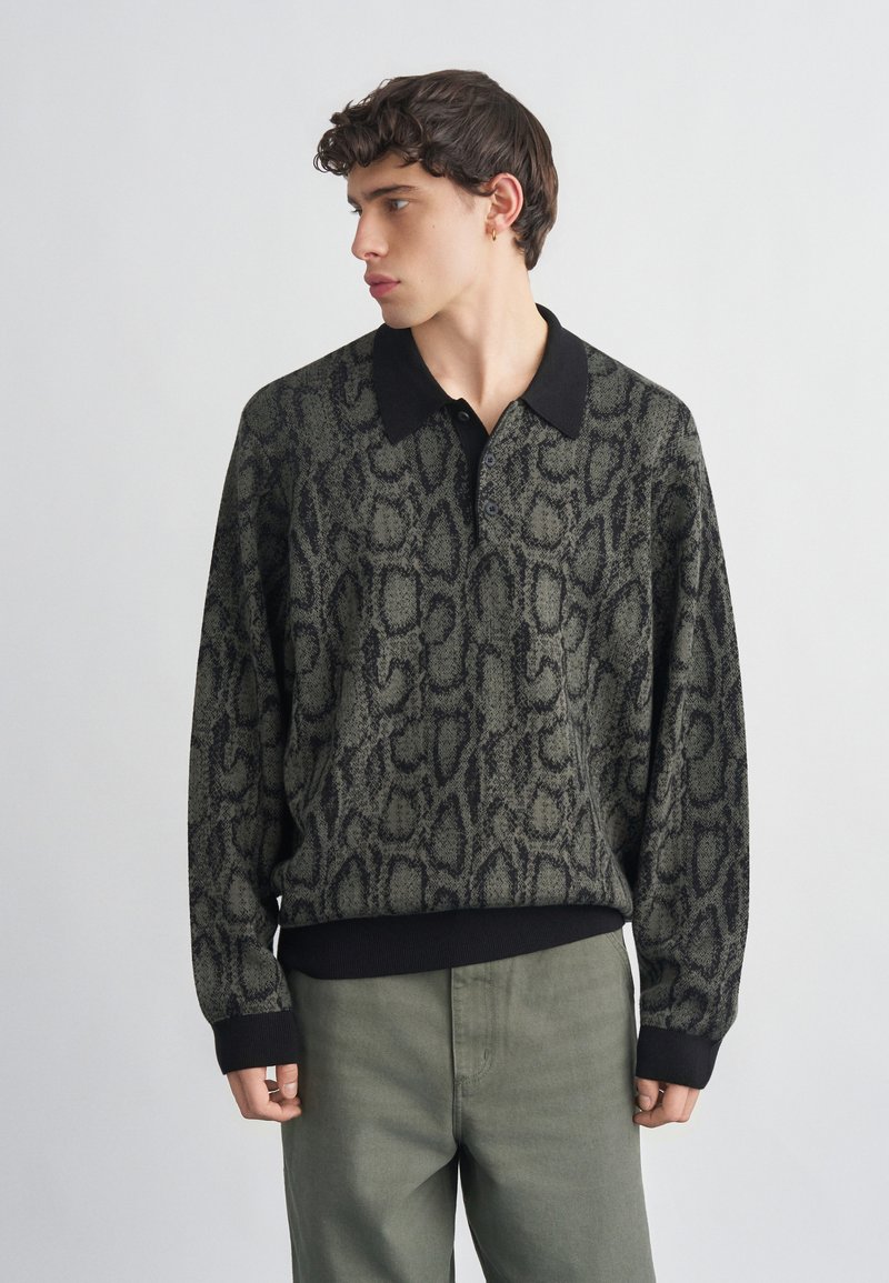 Νέος άνδρας που κοιτάζει προς τα αριστερά, φορώντας ένα μακρυμάνικο πουκάμισο με κουμπιά, μαύρο γιακά και γκρι τύπωμα φιδιού, σε συνδυασμό με πράσινο χακί παντελόνι.