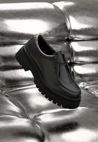 Schwarzer Lederschuh mit einer klobigen Sohle, der eine abgerundete Zehenpartie und ein minimalistisches Schnürdetail aufweist, vor einem glänzenden silbernen Hintergrund.