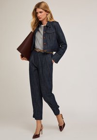 Chaqueta de mezclilla corta con botones grandes, camisa a rayas, pantalones oscuros ajustados azules, cinturón marrón y tacones burdeos, llevando un bolso marrón.