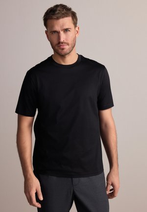 Homme aux cheveux courts bruns et à la barbe, portant un t-shirt noir uni à manches courtes et un pantalon gris foncé sur un fond neutre.