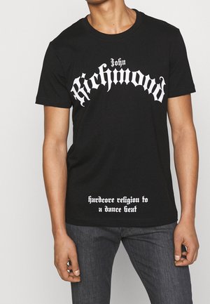 Persoon draagt een zwart t-shirt met "John Richmond" en "hardcore religion to a dance beat", gedrukt in vet witte gotische letters.