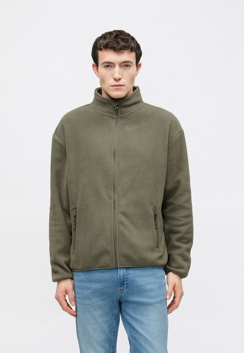 Άνδρας φορώντας φλις ζακέτα με φερμουάρ χρώματος ελιάς και ανοιχτό γαλάζιο τζιν, στέκεται μπροστά σε απλό φωτεινό φόντο.