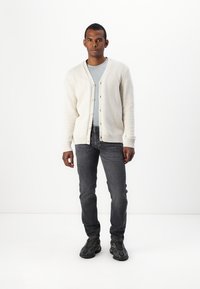Cárdigan de punto beige claro con un patrón texturizado, con botones en la parte delantera, usado sobre una camisa azul claro y jeans gris oscuro, combinado con zapatos negros.