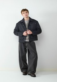 Zwarte geruite jas met een kraag, knoopsluiting en twee voorzakken, gecombineerd met losse grijze broek en donkere schoenen.