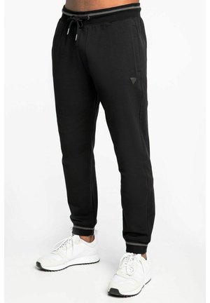Hombre con pantalones deportivos negros con cintura de cordón y zapatillas blancas, de pie sobre un fondo blanco liso.