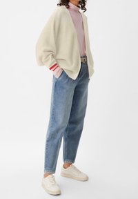 Žena oblečená v krémovém kardiganu přes růžový rolák, světle modrých džínech s vysokým pasem, béžovém pásku a bílých teniskách, stojící s rukama v kapsách.