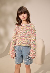 Pull en tricot avec un motif multicolore et manches longues, associé à un short en denim délavé orné de broderies florales. Objet en arrière-plan arrondi.