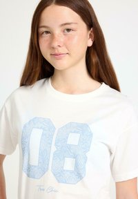Hvid bomulds t-shirt med lyseblå blondemønstrede tal "08" på forsiden og teksten "Tres Chic" under designet.