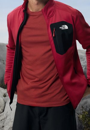 Mężczyzna w czerwonej kurtce zapinanej na zamek błyskawiczny z czarną, zamykaną kieszenią na piersi oraz w rdzawej koszuli, stojący na zewnątrz przy skałach.