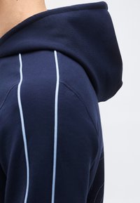 Κοντινό πλάνο στον ώμο ενός σκούρου μπλε φούτερ με κουκούλα, με δύο κάθετες λευκές ρίγες και λεπτομερή ραφή στη ραφή.