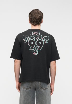 Άνδρας που φοράει μαύρο φαρδύ T-shirt με μεγάλο στυλιζαρισμένο αριθμό «99» και σχεδιασμό σε μορφή φλόγας σε μπλε και ροζ αποχρώσεις στην πλάτη, συνδυασμένο με ξεβαμμένο γκρι τζιν.