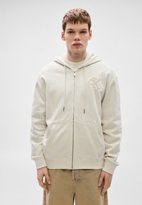 Krém színű cipzáros kapucnis pulóver elülső zsebbel, húzózsinóros kapucnival és hímzett logóval. Puha anyagból készült, lazított szabással és bordázott mandzsettával.