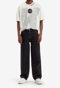 Persona con una camiseta gráfica blanca de gran tamaño, jeans negros holgados y zapatillas blancas, de pie contra un fondo blanco liso.