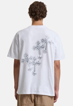 Personne aux cheveux courts et bouclés porte un t-shirt blanc avec quatre croix argentées au dos, associé à un pantalon beige.