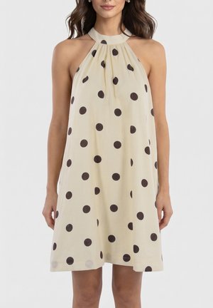 Femme portant une robe crème sans manches à col licou avec de grands pois noirs, debout devant un fond clair uni.