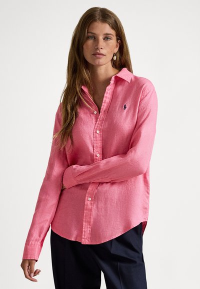 Sieviete ar gariem brūniem matiem, kas valkā rozā pogājamu kreklu un tumšas bikses, stāv ar vienu roku pār vidukli uz baltas fona.