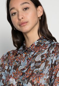 Camisa com estampado floral, apresentando um fundo castanho, padrões coloridos de paisley e flores, design com botões e colarinho pontudo.