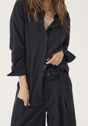 Kobieta ubrana w czarną koszulę w drobną kratkę na guziki i pasujące szerokie spodnie z paskiem, ręce poprawiają koszulę i okolice talii.