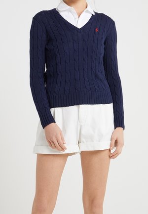 Granatowy sweter w splot warkoczowy z dekoltem w serek, noszony na białej koszuli z kołnierzykiem, zestawiony z białymi podwiniętymi szortami, pokazujący górną część ciała i nogi.
