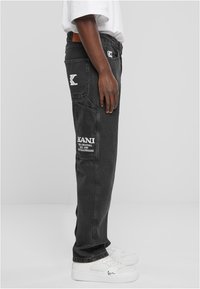 Pantaloni cargo in denim nero con tasche laterali; presenta il logo e il branding KANI. Indossati con sneaker bianche. Taglio dritto e tessuto robusto.