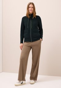 Tmavě zelená bunda na zip s dvěma předními kapsami, strukturovaná látka, stažený límec, kombinovaná s béžovými kalhotami s širokými nohavicemi a bílými teniskami.